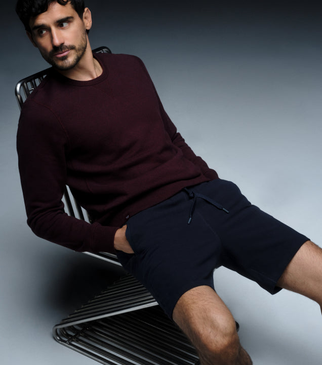 image of a man wearing a maroon long sleeve shirt and navy blue ace sweatshorts, lounging on a metal chair