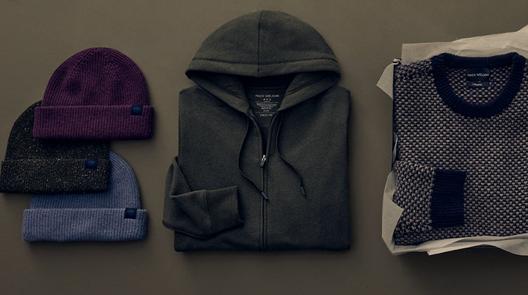 A styled flat-lay of various men's clothing items, including a dark gray hoodie, a purple knit beanie, a blue plaid shirt, a gray t-shirt, and folded garments, arranged on a dark gray background.